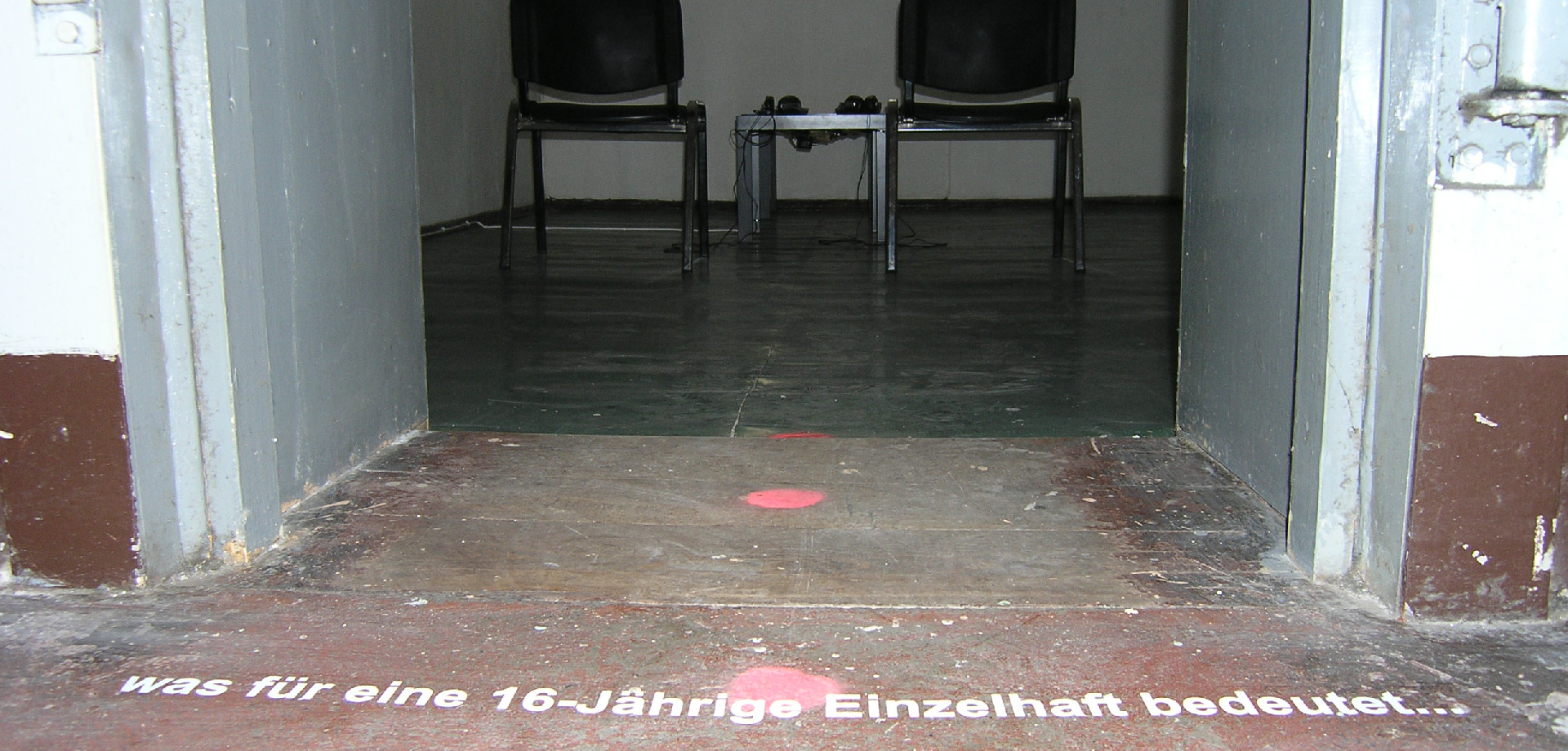 zwei Stühle stehen in einem Raum, vor dem Raum auf dem Boden steht "Was für eine 16jährige Einzelhaft bedeutet..."