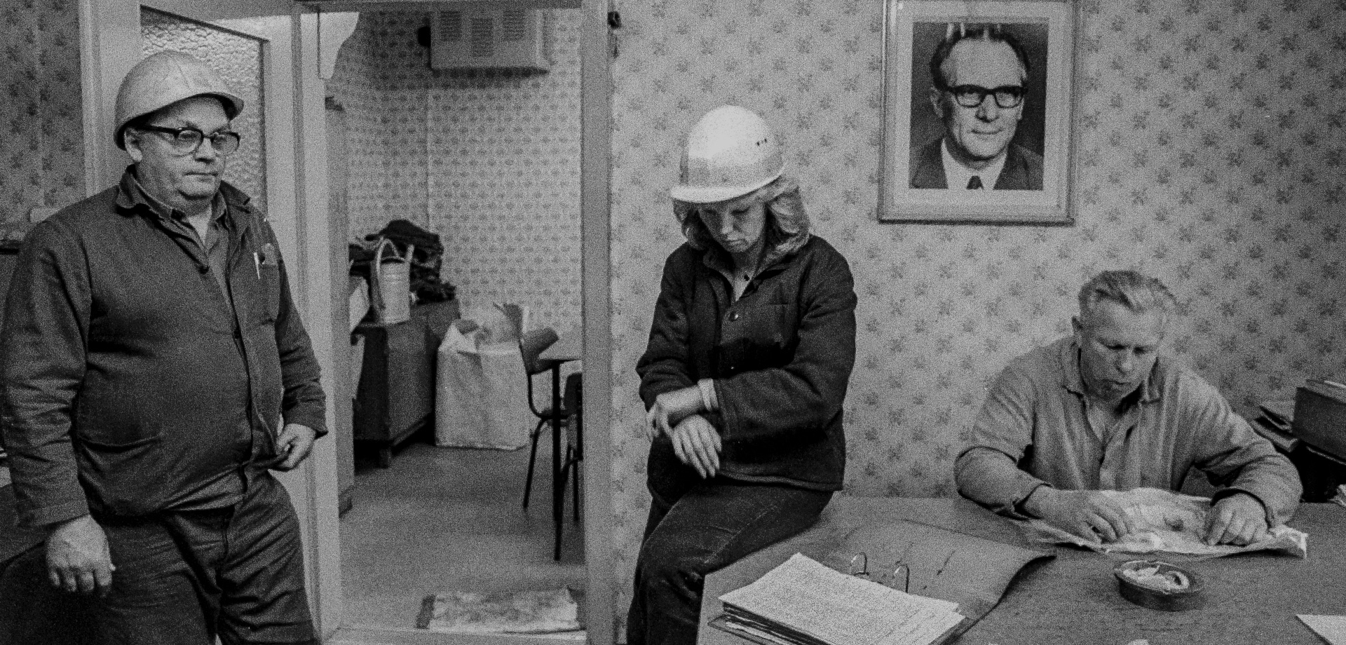 zwei Männer und eine Frau sind in einem Büro, ein Mann und die Frau tragen Sicherheitshelme, an einer Wand hängt ein Porträt von Erich Honecker