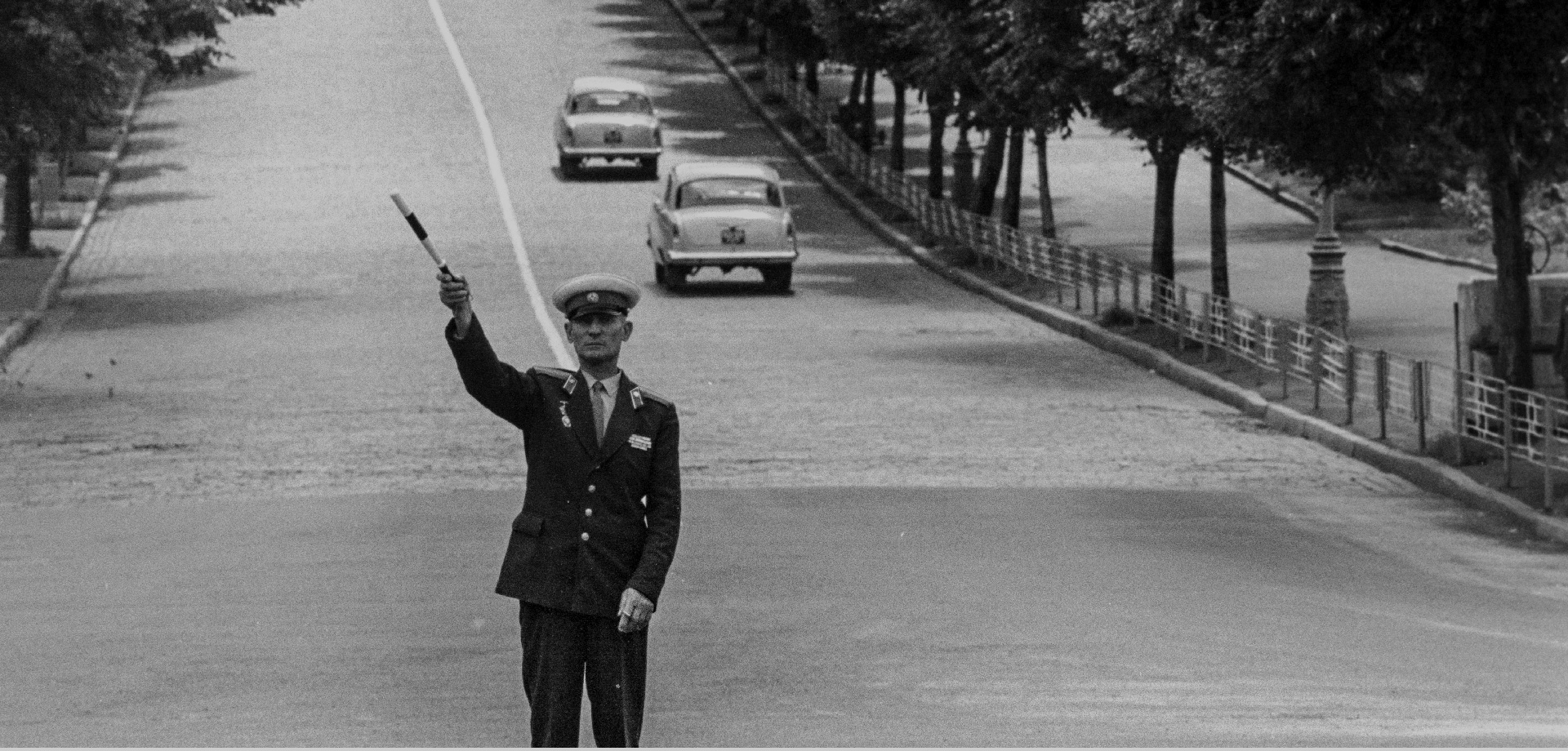 ein Polizist steht auf einer wenig befahreren Straße, er hebt einen Stab, zwei Wartburgs fahren hinter ihm